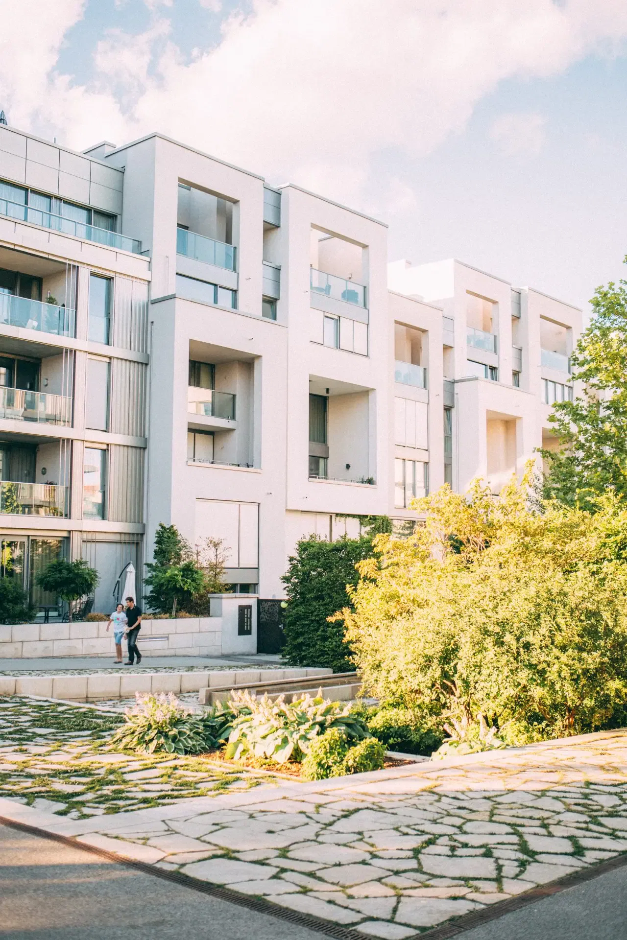 Modern white apartment building with large balconies, lush green landscaping in front, and two people walking on the sidewalk.