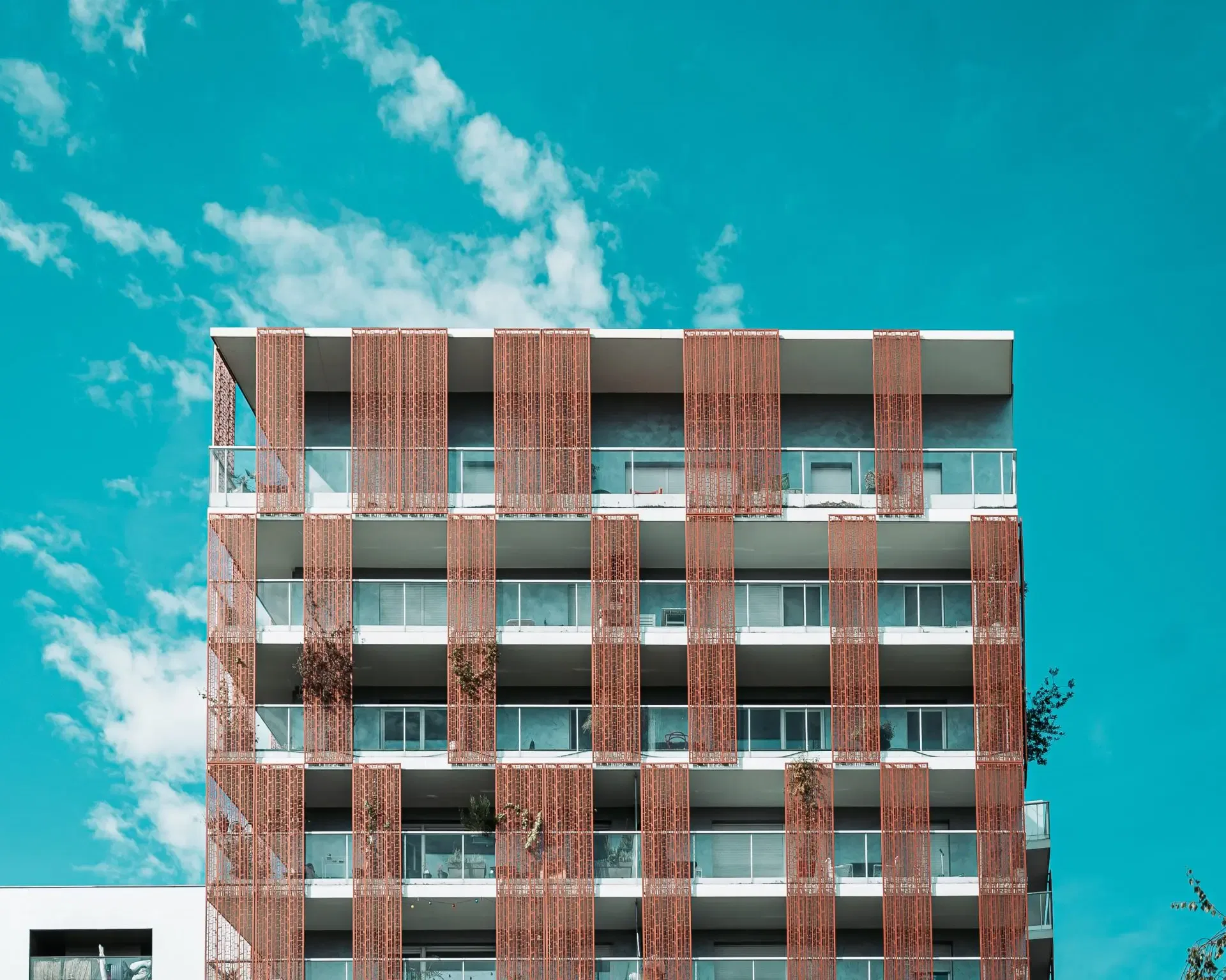 Modern apartment building with glass balconies and red vertical slats on the facade, set against a bright blue sky with scattered clouds.