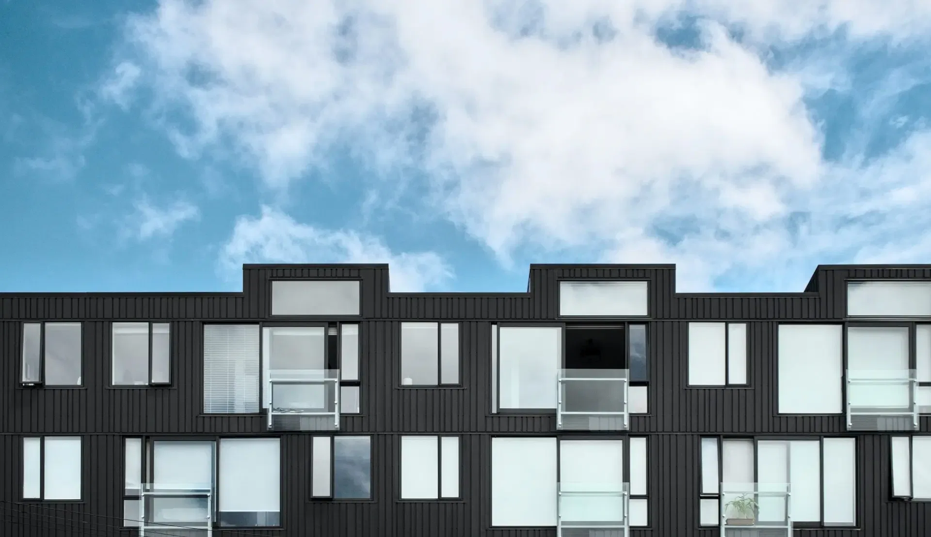 Modern black apartment building with large windows and glass balconies under a partly cloudy blue sky.
