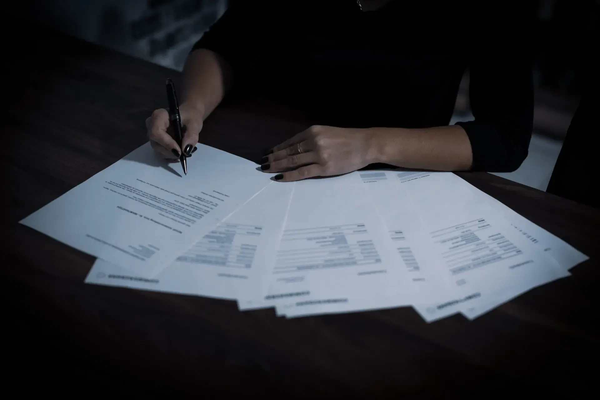 Person with black-painted nails signing multiple documents on a dark wooden table.