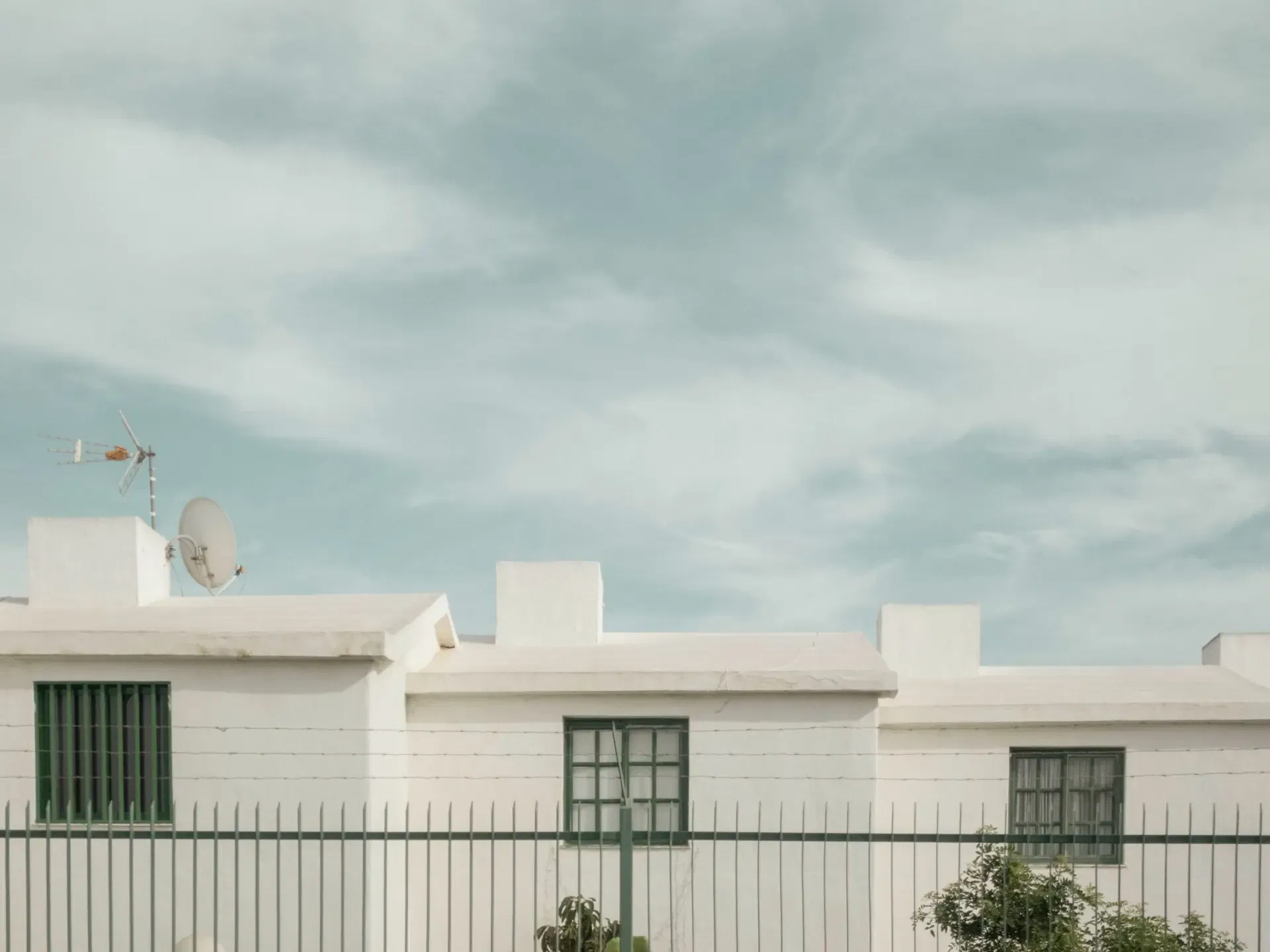 White building with green windows and a fence in the foreground under a cloudy sky, featuring an antenna and satellite dish.
