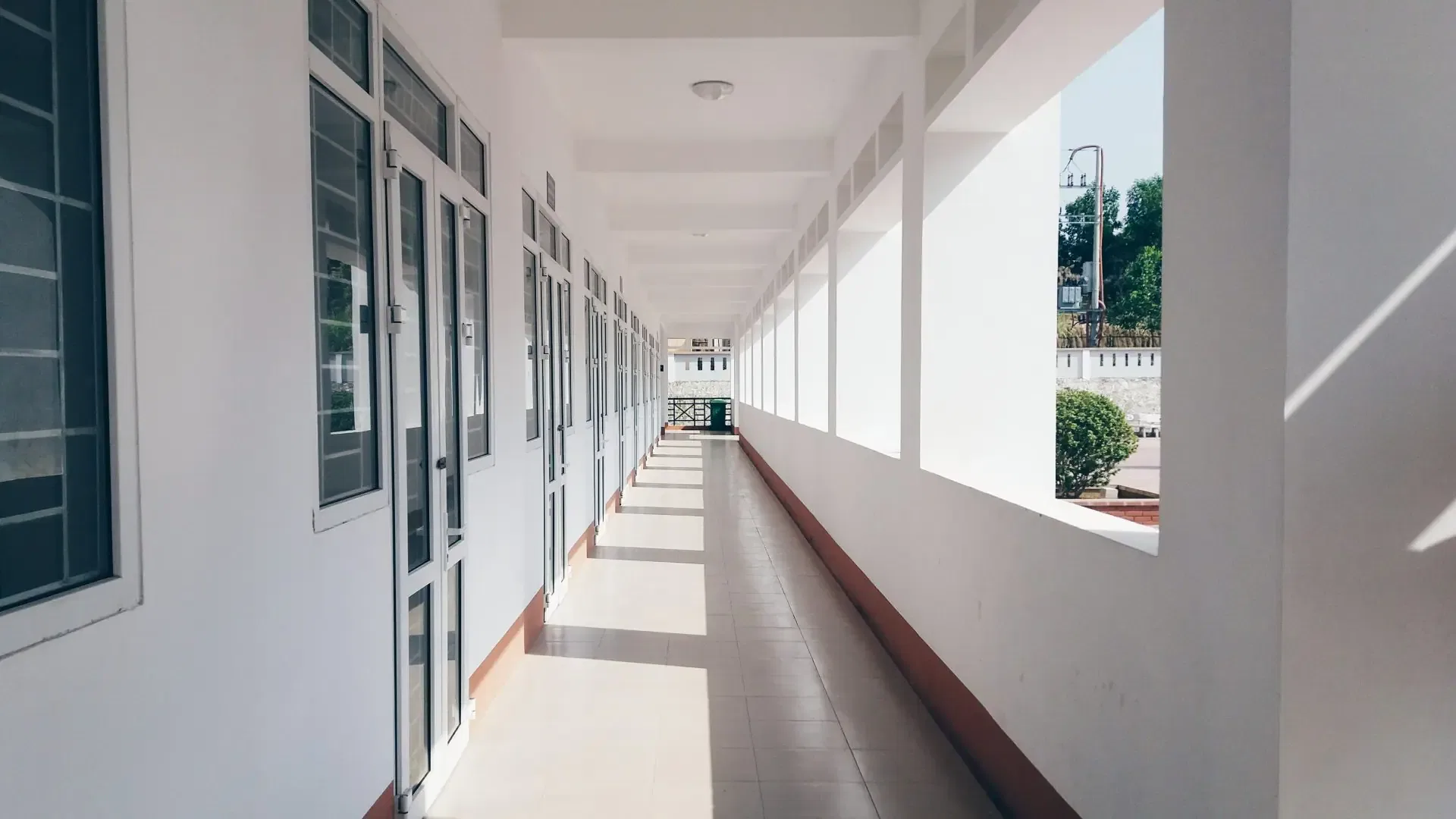 Sunny, empty hallway with white walls and doors on the left. Large windows on the right overlook a courtyard with greenery.