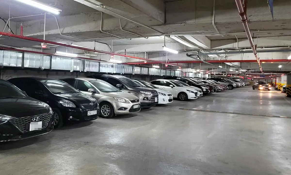 Cars are parked in rows in a well-lit, underground parking garage with concrete ceilings and red pipes.