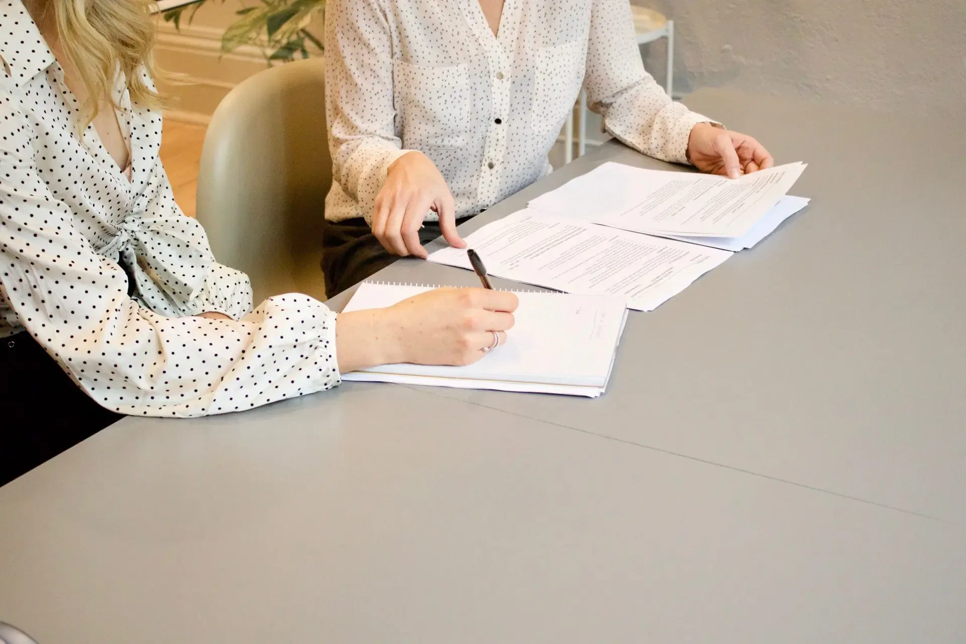 Two people discuss documents at a table, one writing in a notebook and the other pointing to papers.