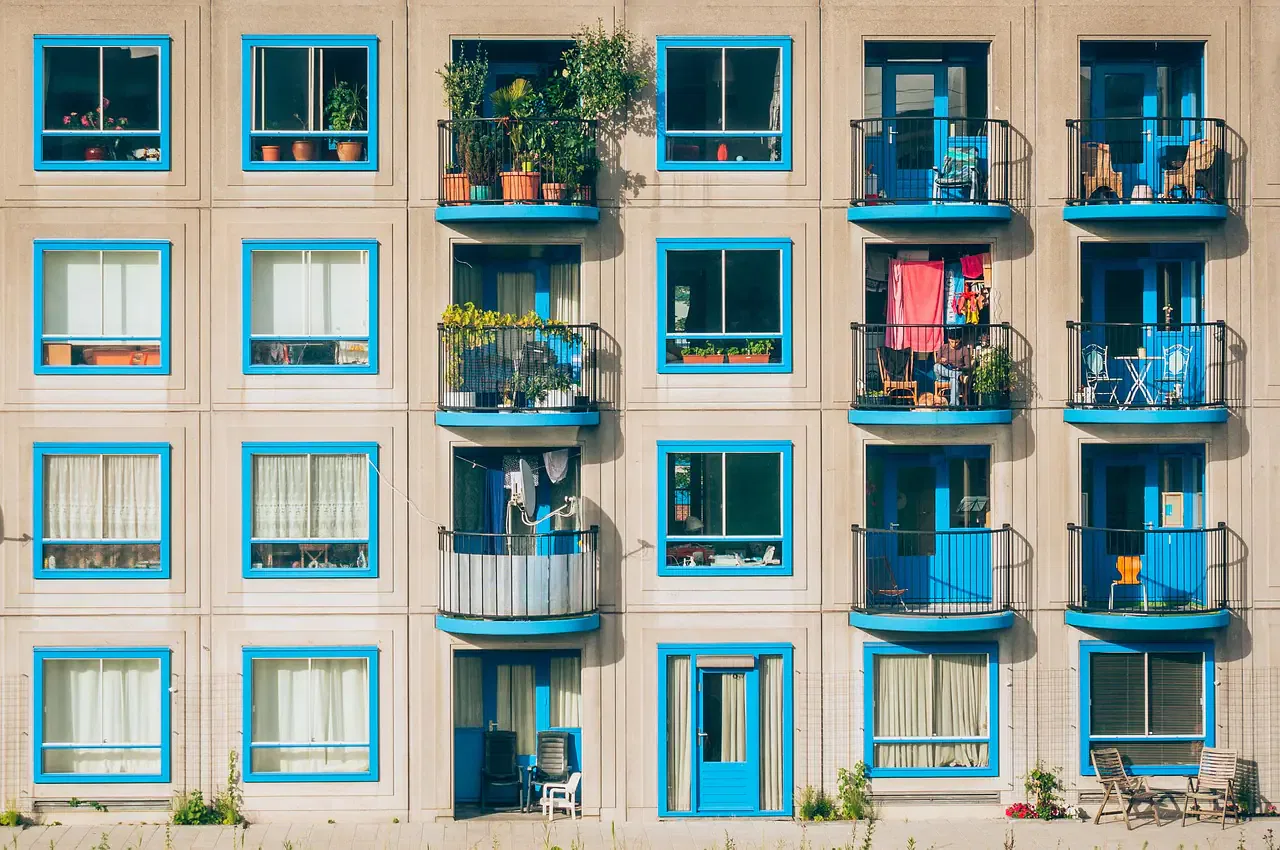 Apartment building with blue-framed windows and balconies, some with plants and laundry hanging, viewed on a sunny day.