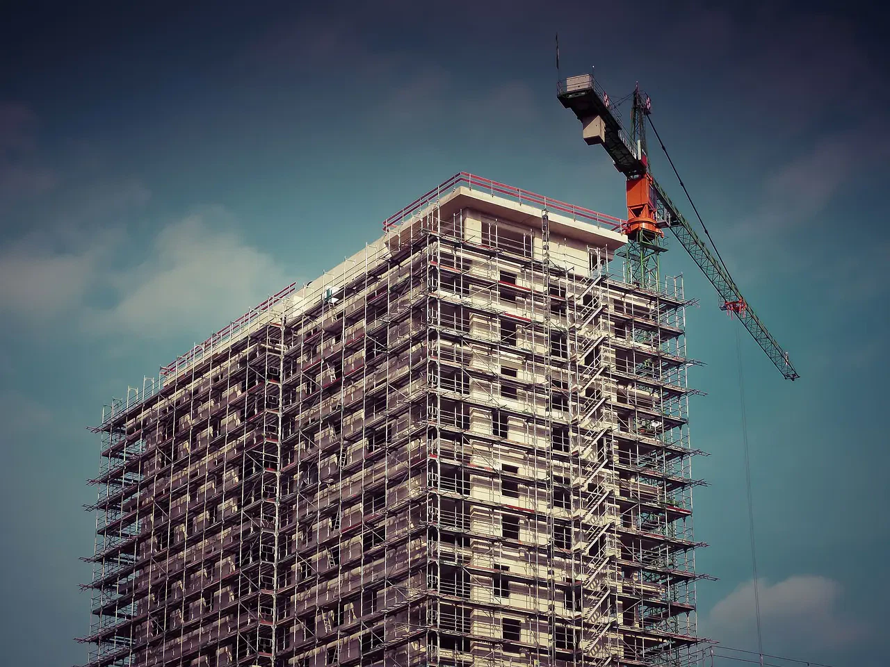 Tall building under construction with scaffolding and a large crane against a cloudy sky.