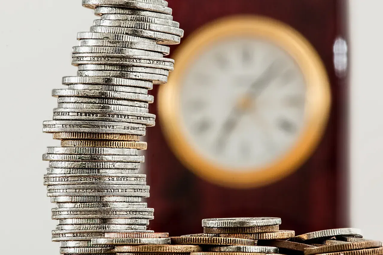 Stacks of silver coins in focus with a blurred vintage clock in the background.