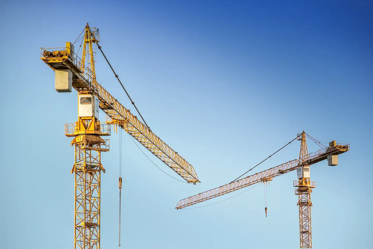 Two yellow construction cranes against a clear blue sky, positioned with one slightly in front of the other.