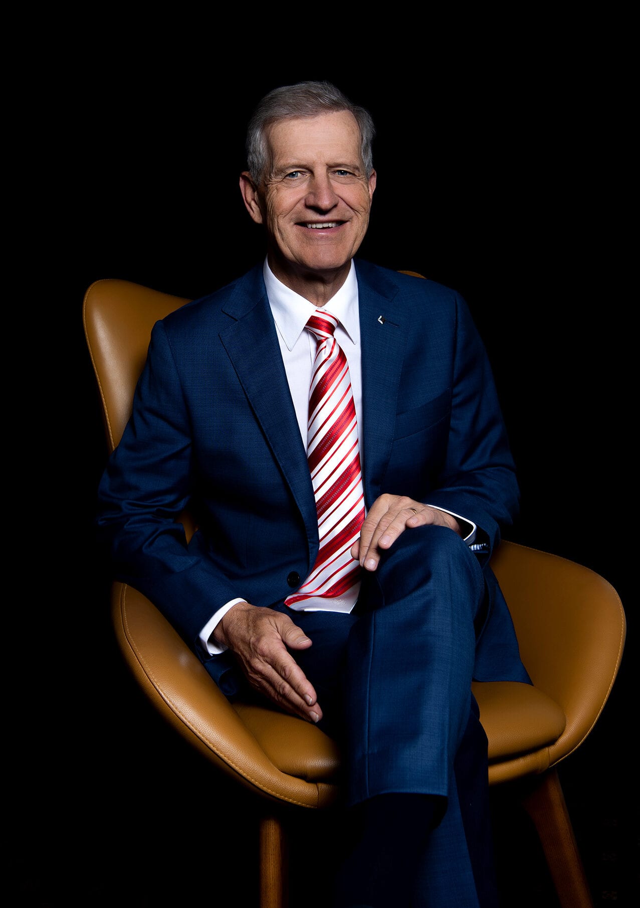 Smiling senior businessman in blue suit and red tie sits in a brown leather chair.