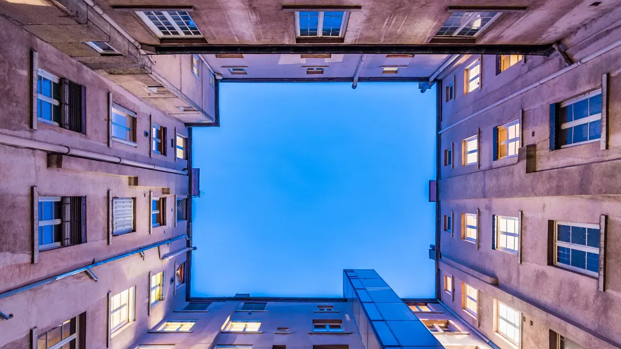 Looking up at a blue sky surrounded by four walls of an apartment building with lit windows in the evening.