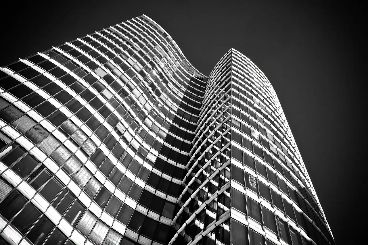 Upward view of a modern skyscraper with a wavy facade, featuring reflective glass windows against a dark sky. Black and white.