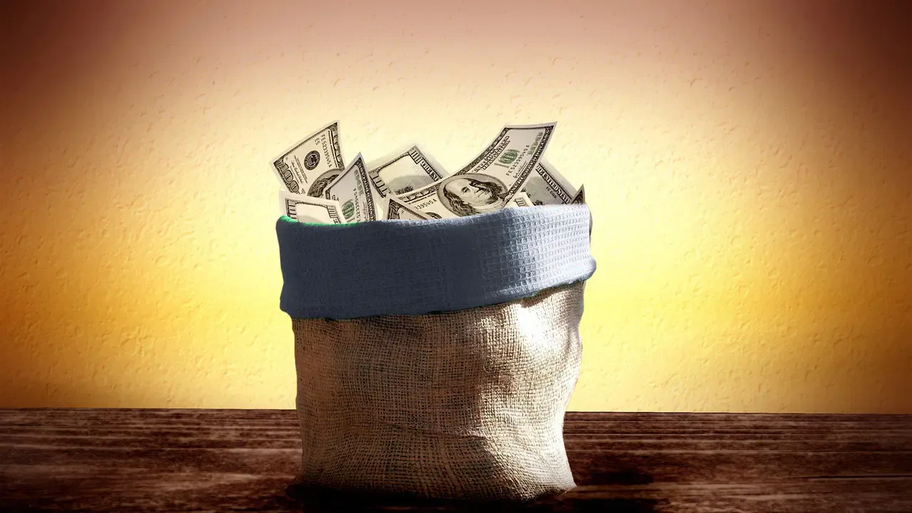 A burlap sack filled with US dollar bills, sitting on a wooden surface against a yellow background.
