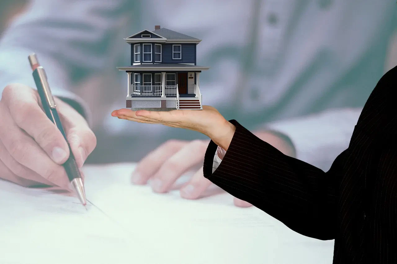 A hand in a business suit holds a small house model, with a person in the background signing documents.