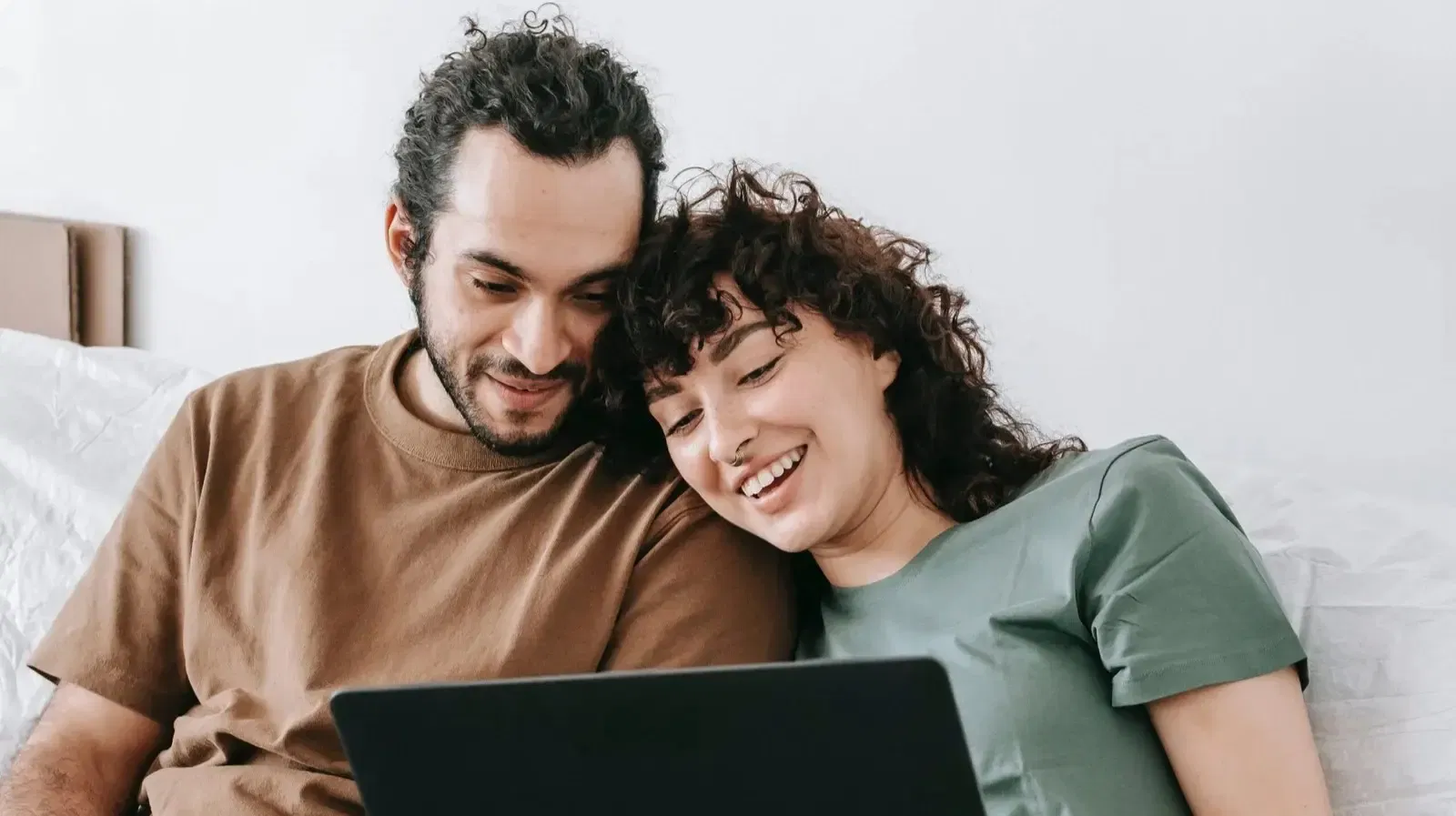 A couple sits on a couch, smiling and looking at a laptop together.