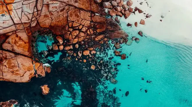 Aerial view of a rocky coastline merging into clear turquoise waters and a white sandy beach.