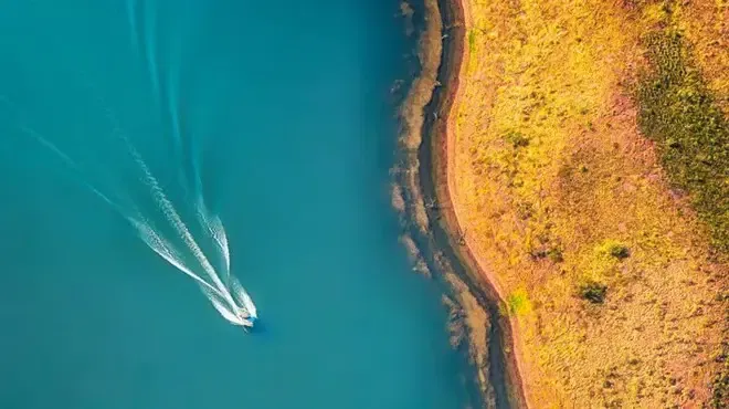 Aerial view of a boat speeding across turquoise water, leaving a white wake, next to a golden, grassy shoreline.