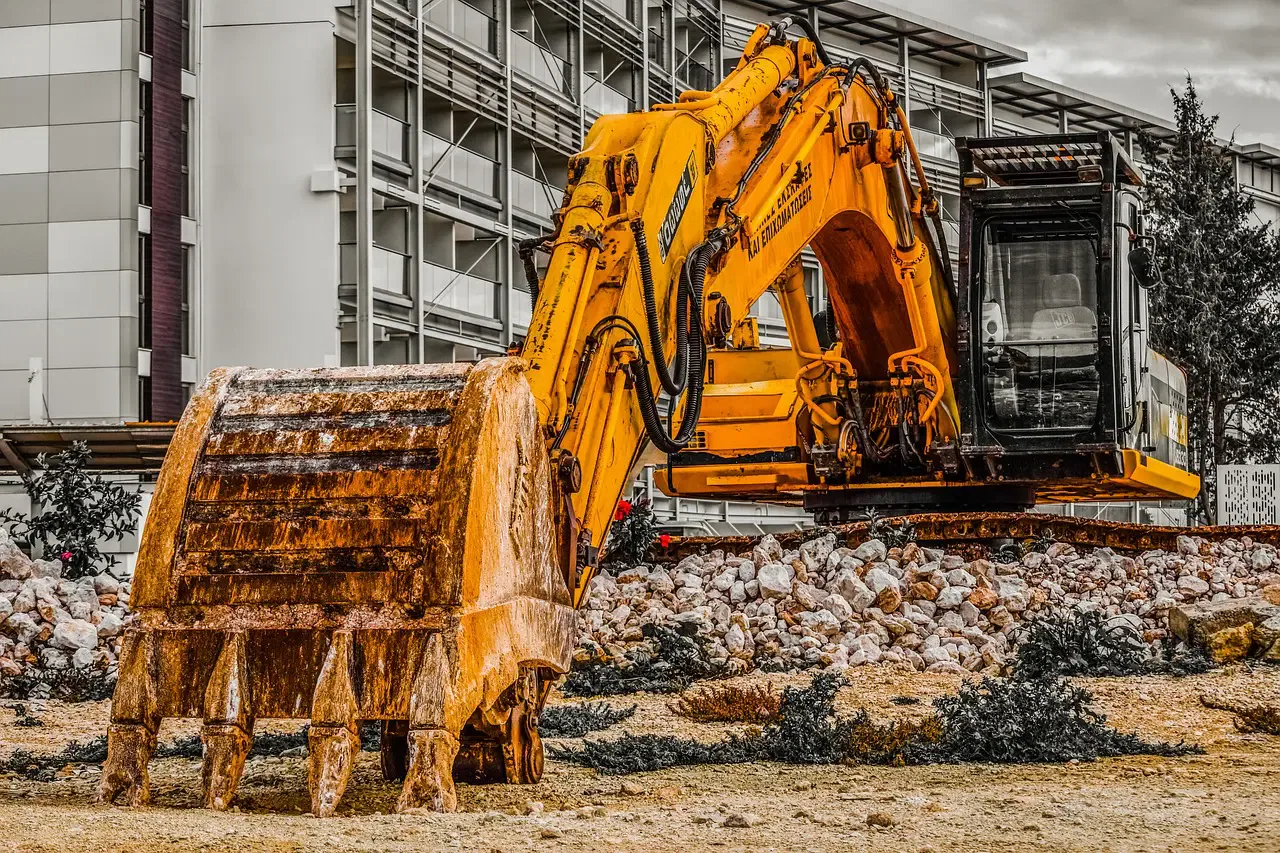 A yellow excavator on a construction site with a building in the background. The bucket is resting on a pile of rocks.
