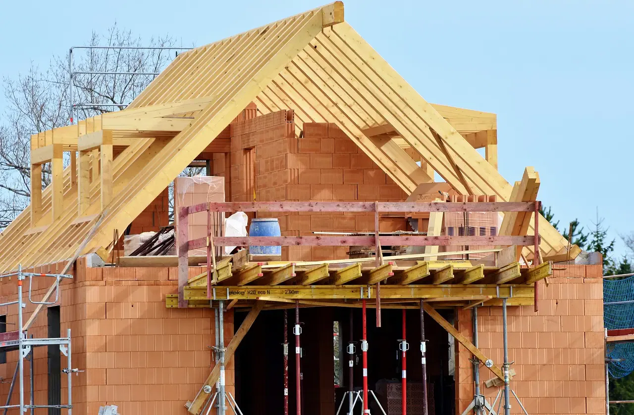 Partially constructed house with a wooden frame and exposed brick walls under a clear sky. Scaffolding and construction materials visible.