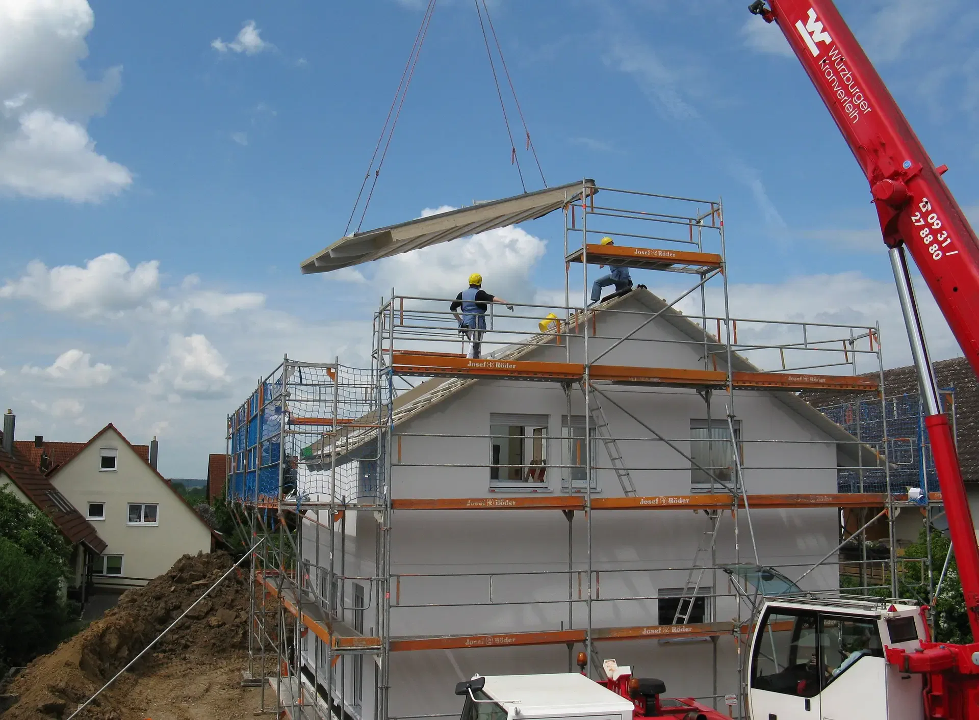 Construction workers on scaffolding install a roof panel on a new house using a crane on a sunny day.