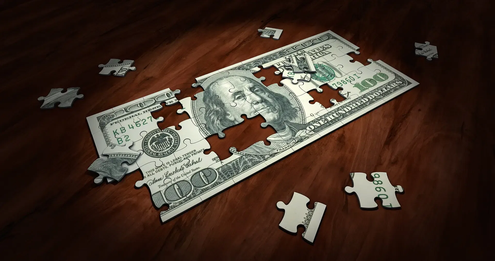 A $100 bill depicted as a jigsaw puzzle with some pieces missing, lying on a wooden surface.