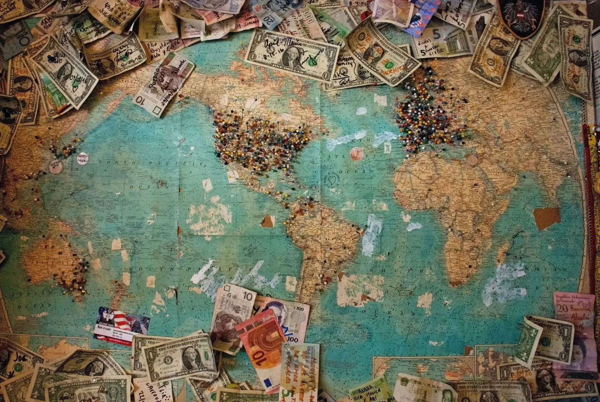 A vintage world map covered with colorful pins, surrounded by various international paper currency.