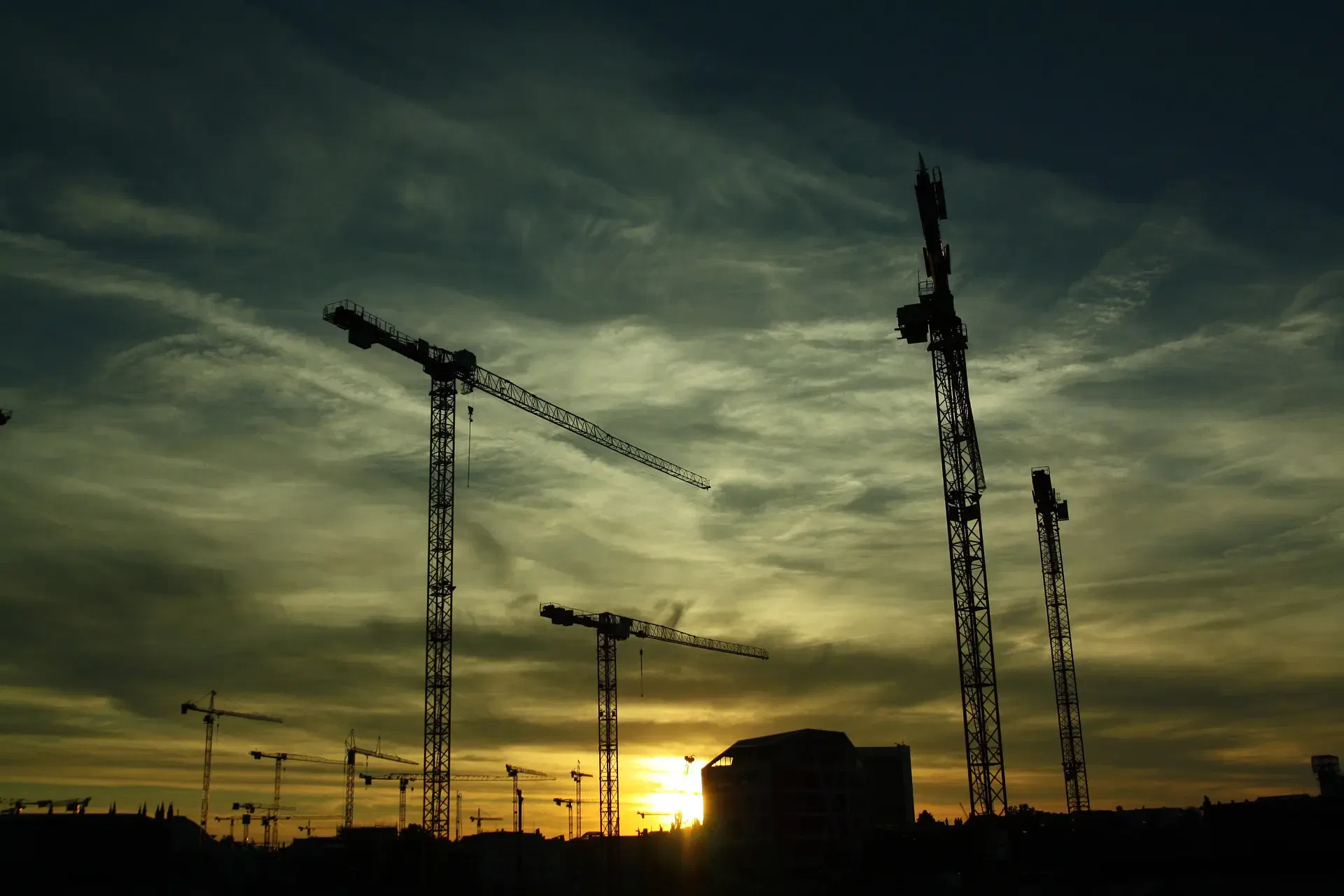 Silhouettes of construction cranes against a dramatic sunset sky, with a partially constructed building in the background.