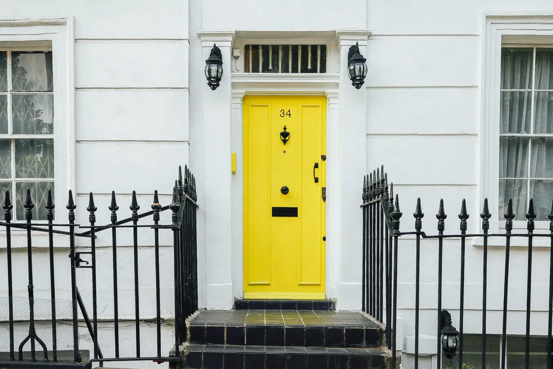 Bright yellow front door with the number 34, surrounded by black iron railings, set in a white facade with two sconces and windows.