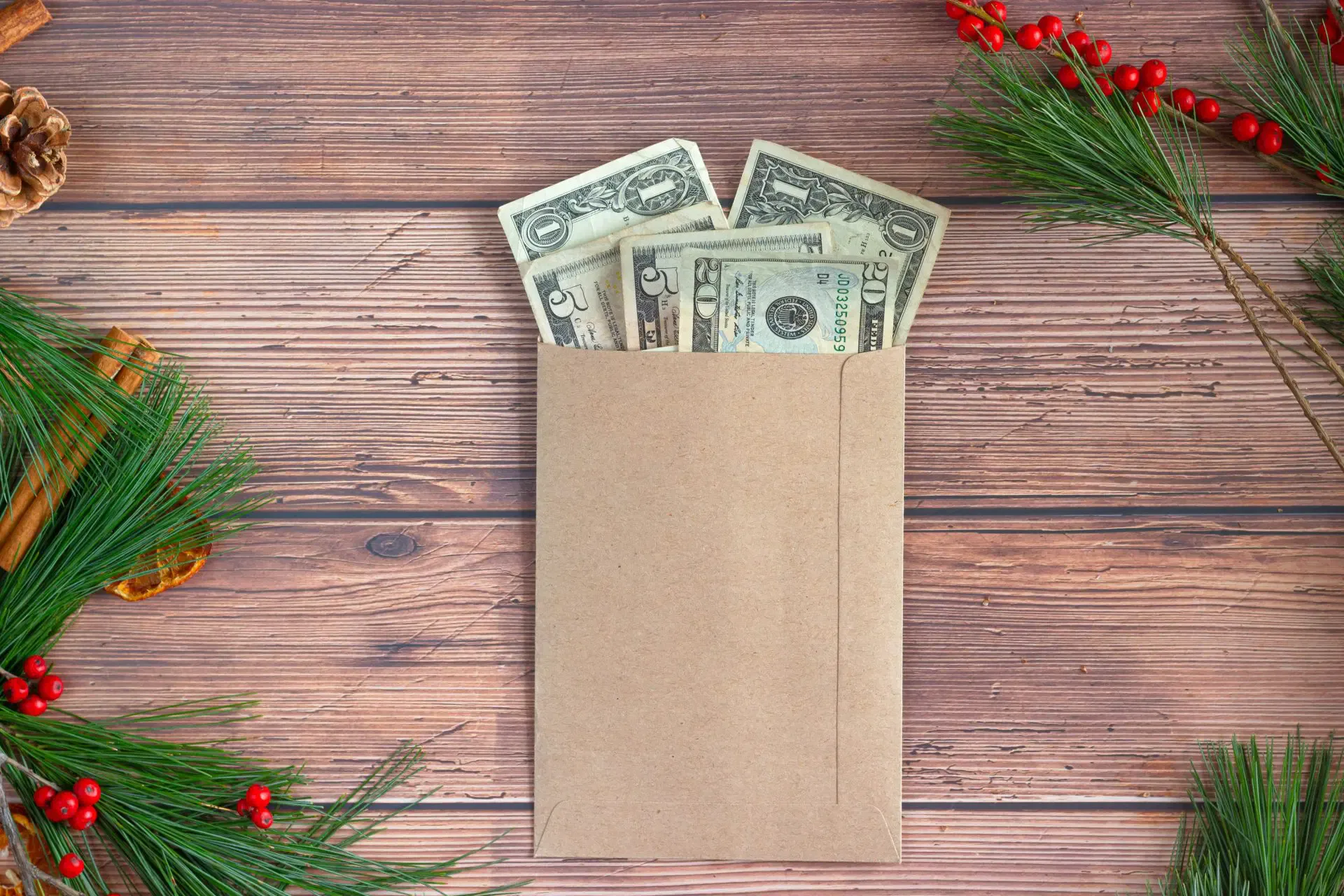Envelope with cash bills on wooden table, surrounded by pine branches, red berries, pinecones, and cinnamon sticks.