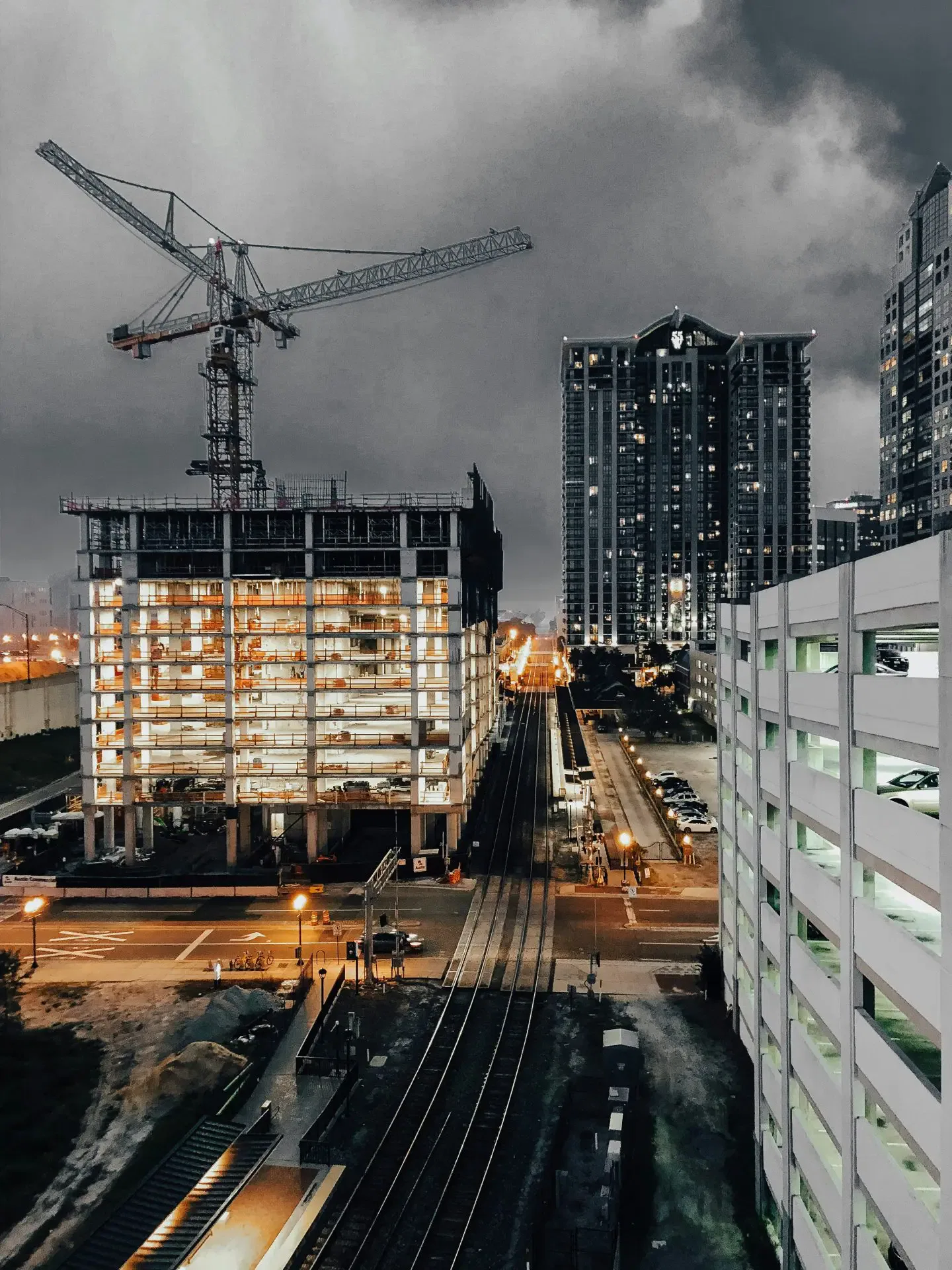 Nighttime cityscape featuring a construction site with a crane, adjacent railway tracks, and lit-up high-rise buildings.