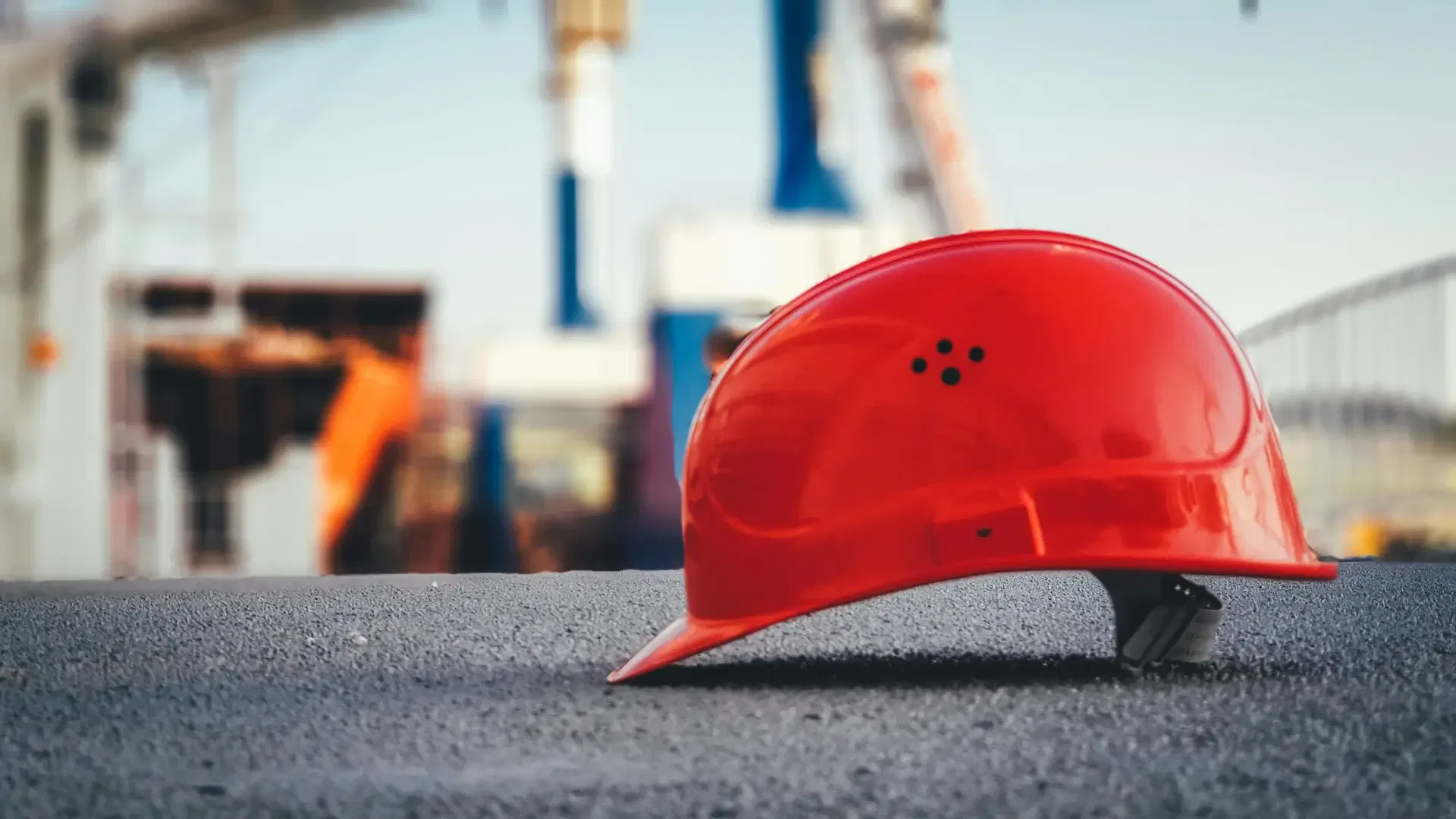 Red construction helmet resting on asphalt, with a blurred construction site background.