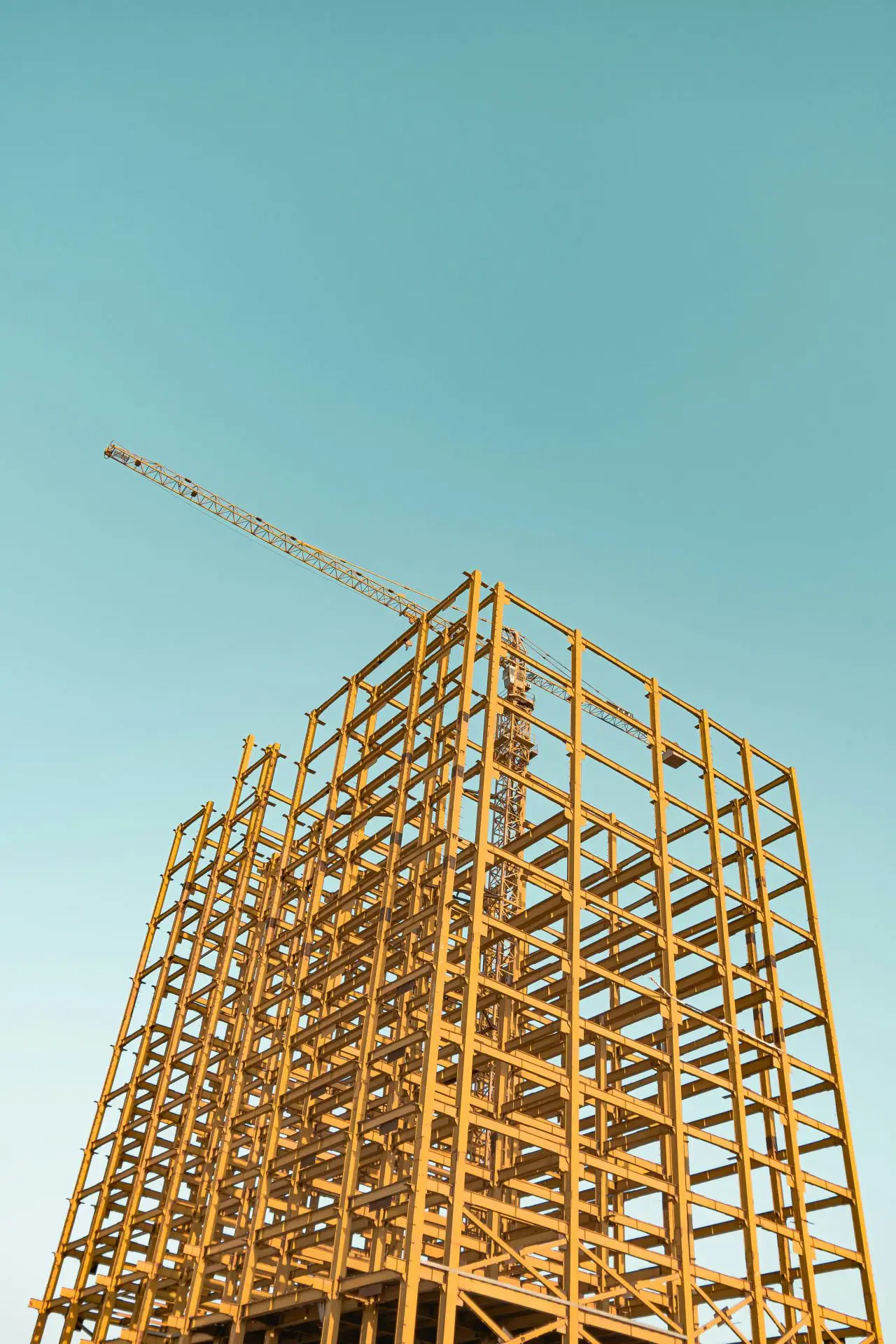 Steel framework of a high-rise building under construction with a crane against a clear blue sky.