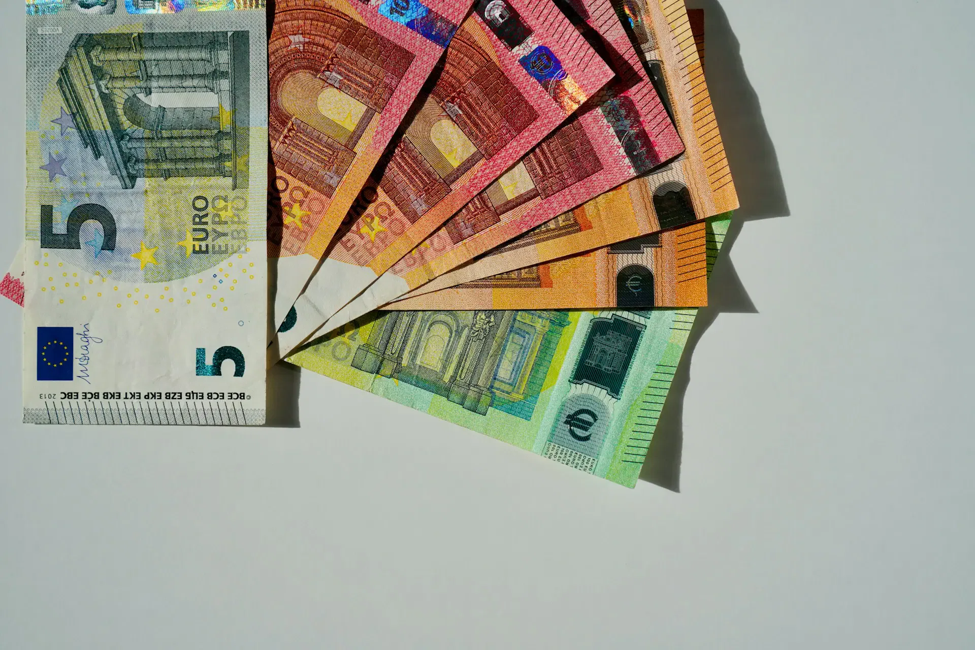 A fan of Euro banknotes, including denominations of 5, 10, 20, 50, 100, and 200, spread out on a white surface.