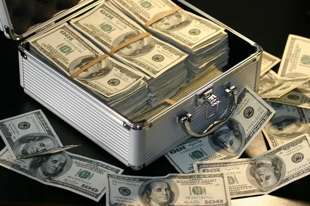 A metallic briefcase filled with neatly stacked 100-dollar bills, surrounded by loose 100-dollar bills on a table.