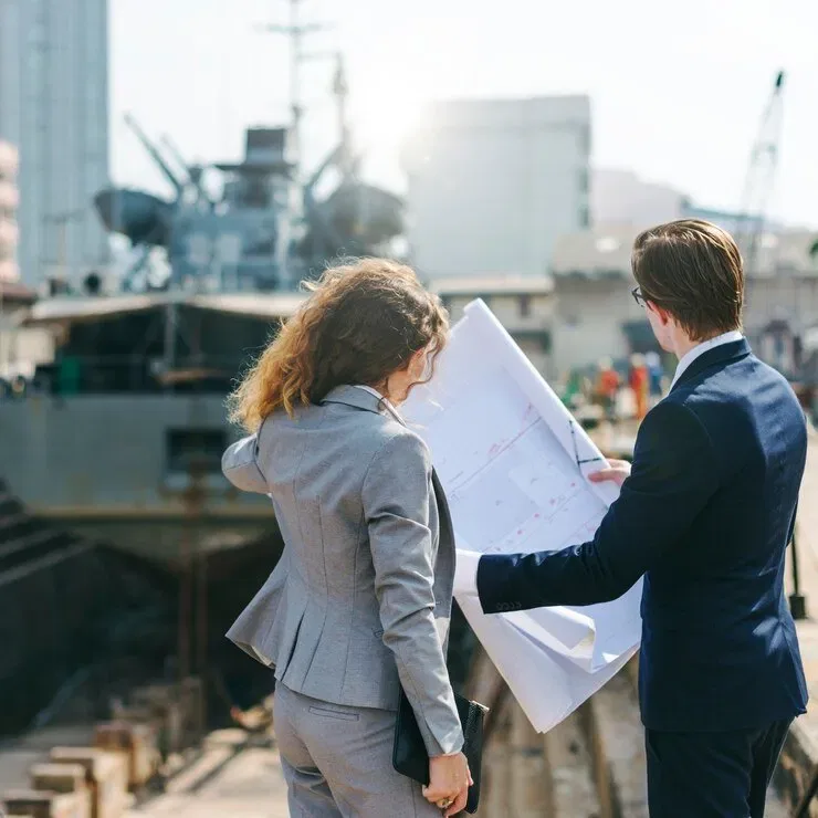 Two businesspeople review blueprints near a ship in a Danish shipyard. Danish estate planning.