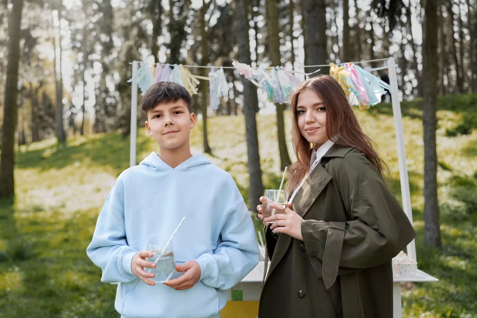 Two young people, a boy and a girl, stand at a lemonade stand in a park.