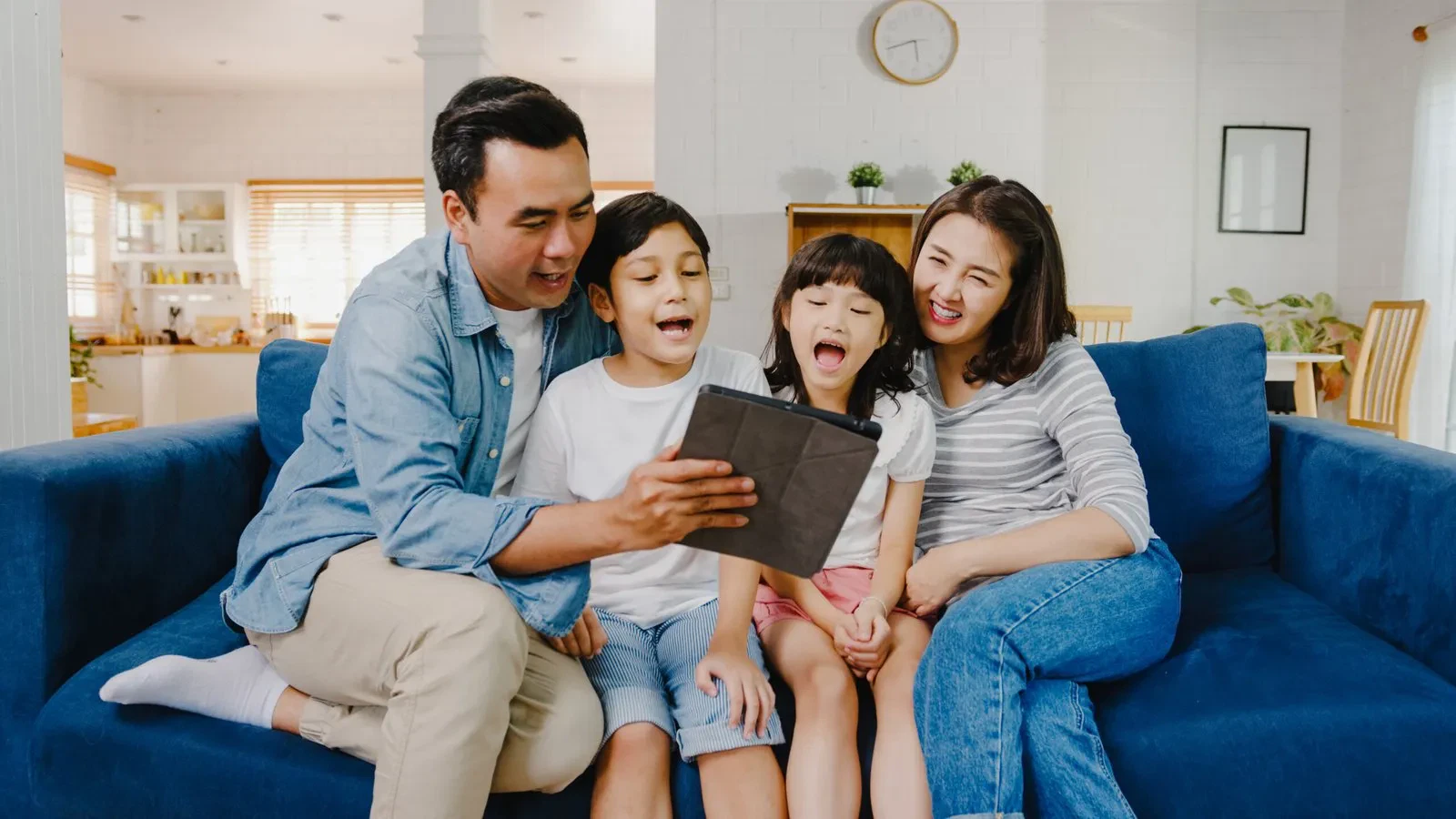 Happy family of four sitting on a blue couch, sharing a tablet at home