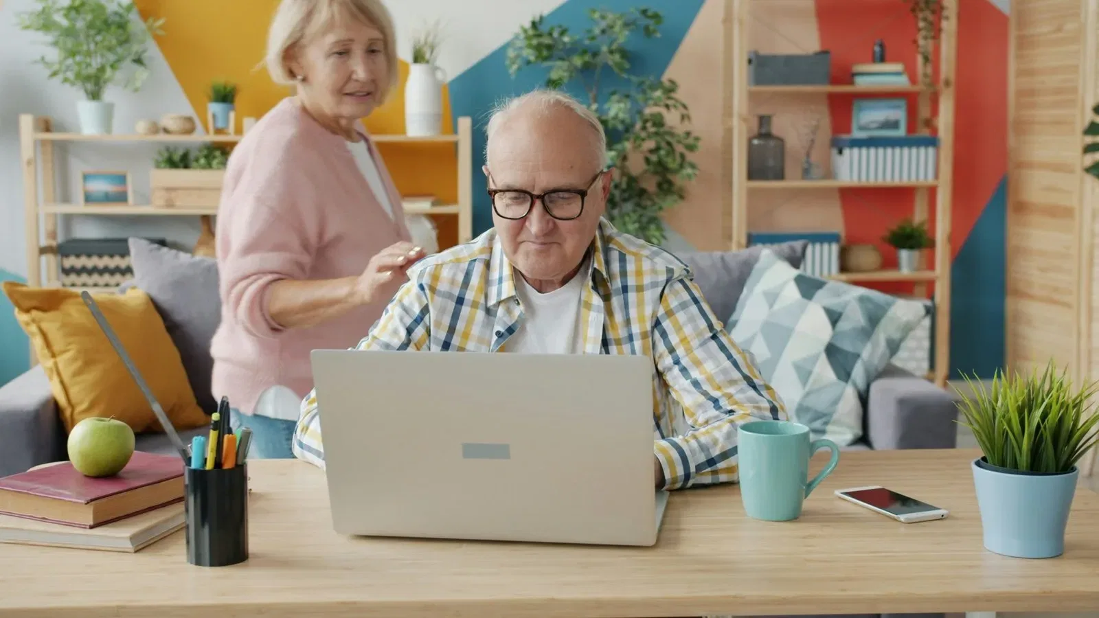 Older man using laptop, woman looking over his shoulder. Home office setting.