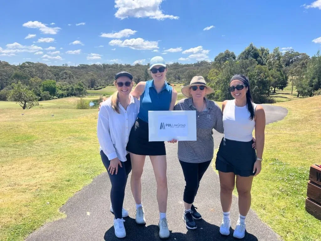 Four women golfers holding a PBL Law Group sign on a sunny course.