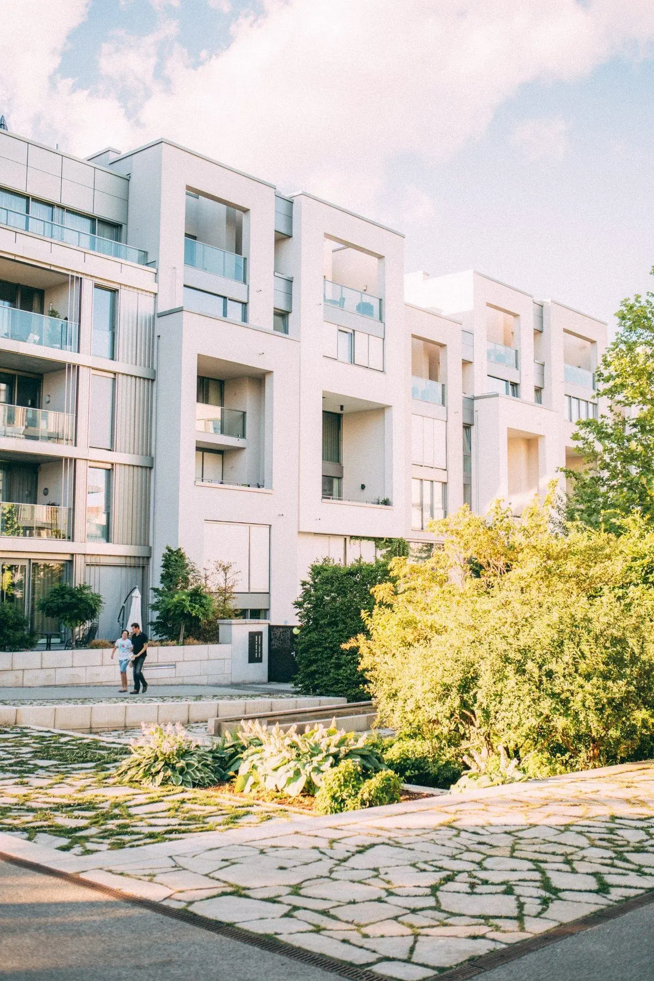 Modern white apartment building with large balconies, lush green landscaping in front, and two people walking on the sidewalk.