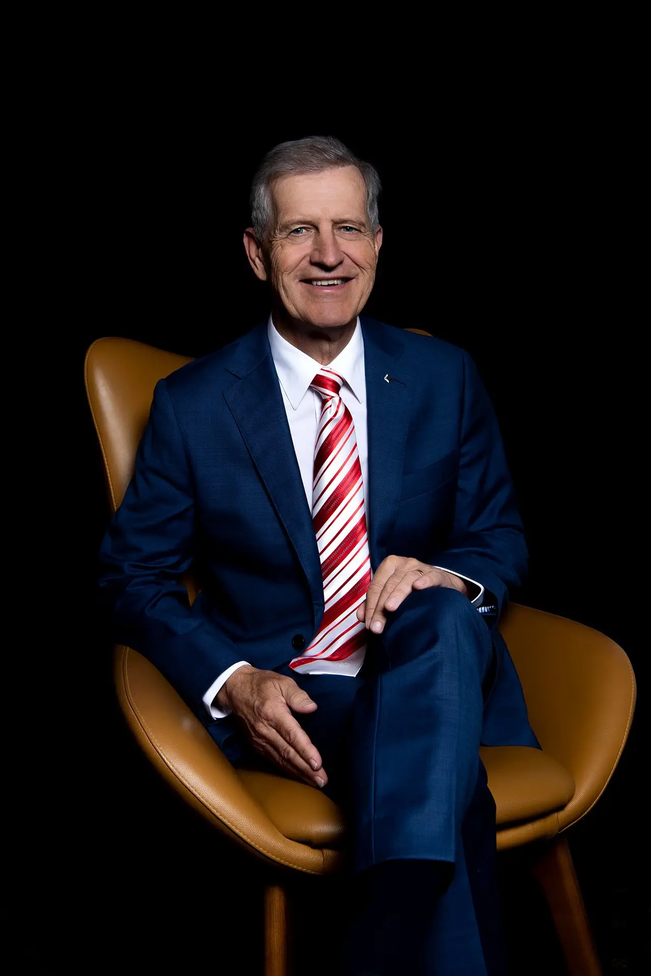 Smiling senior businessman in blue suit and red tie sits in a brown leather chair.