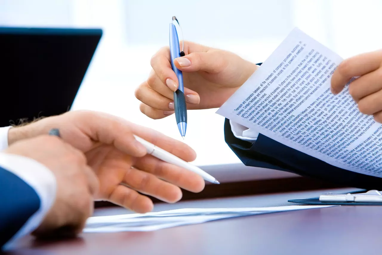 Close-up of two people exchanging pens and reviewing a document with a laptop in the background.