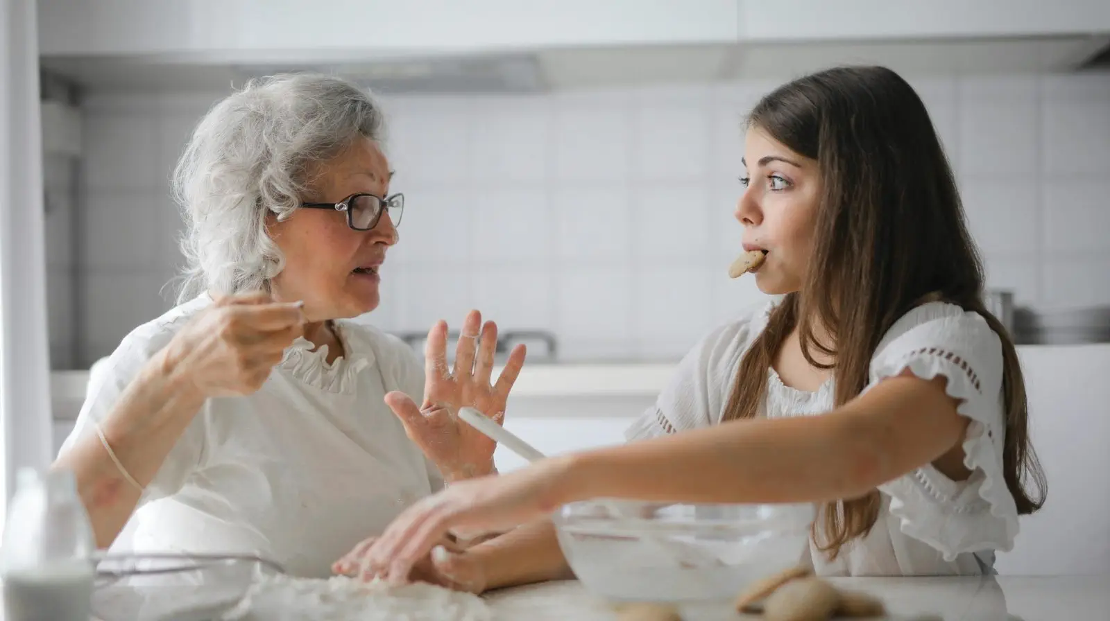 An elderly woman and a young girl bake together in a kitchen; the girl has a cookie in her mouth while listening intently.