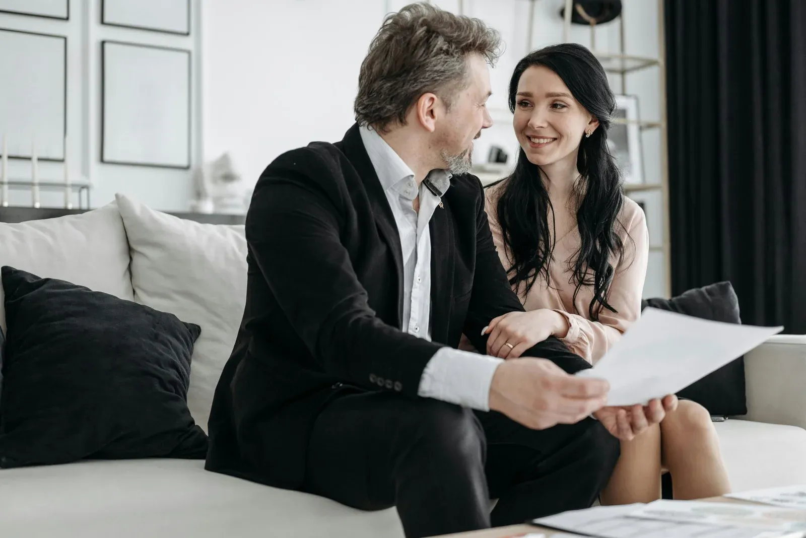 A man and a woman are sitting on a couch, smiling and holding a document, engaged in conversation in a modern living room.