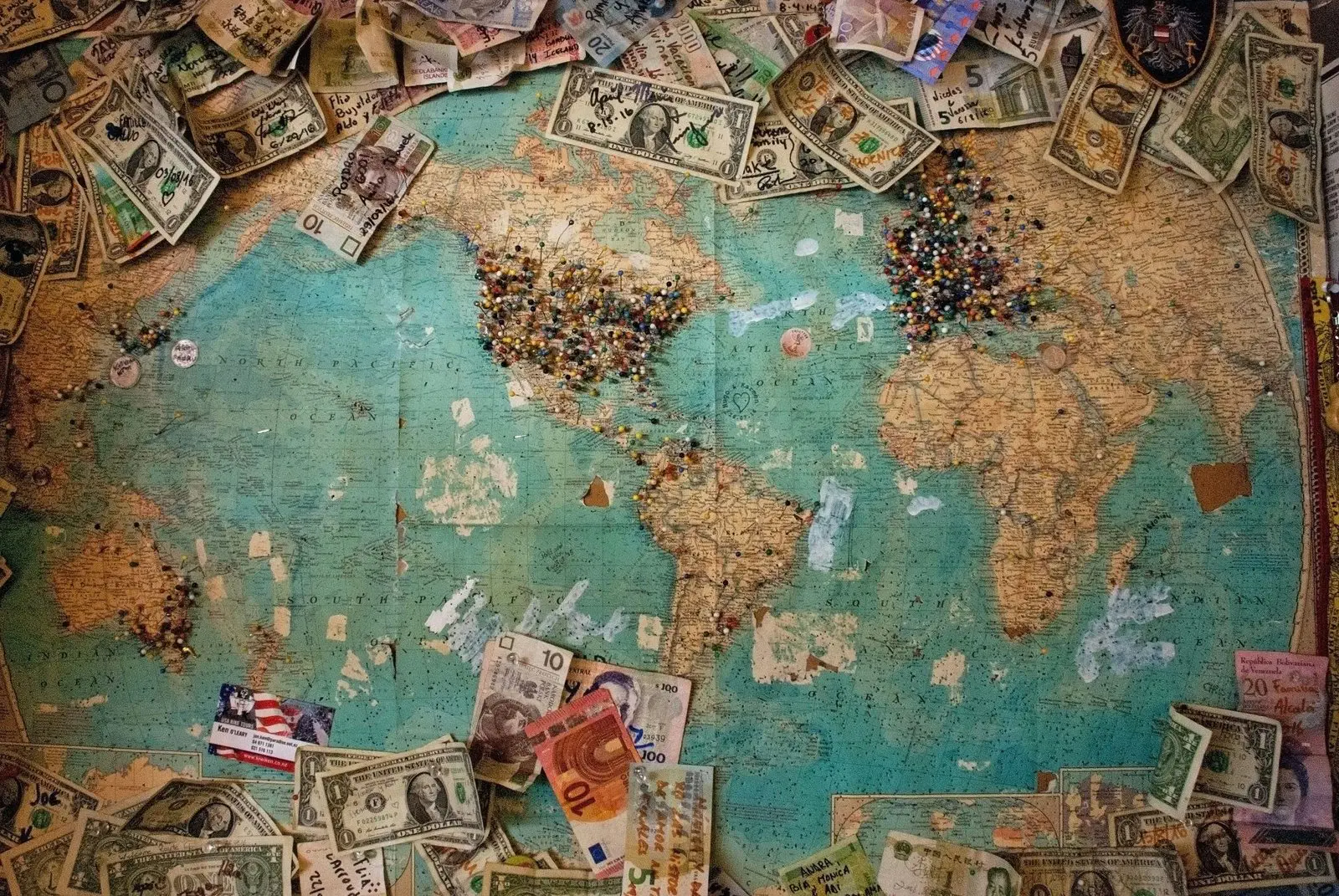 A vintage world map covered with colorful pins, surrounded by various international paper currency.