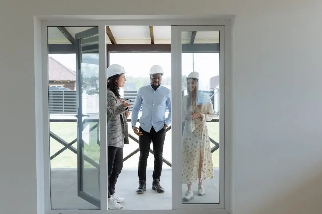 Three people wearing hard hats discuss home building defects and statutory warranties, viewed through a glass door.