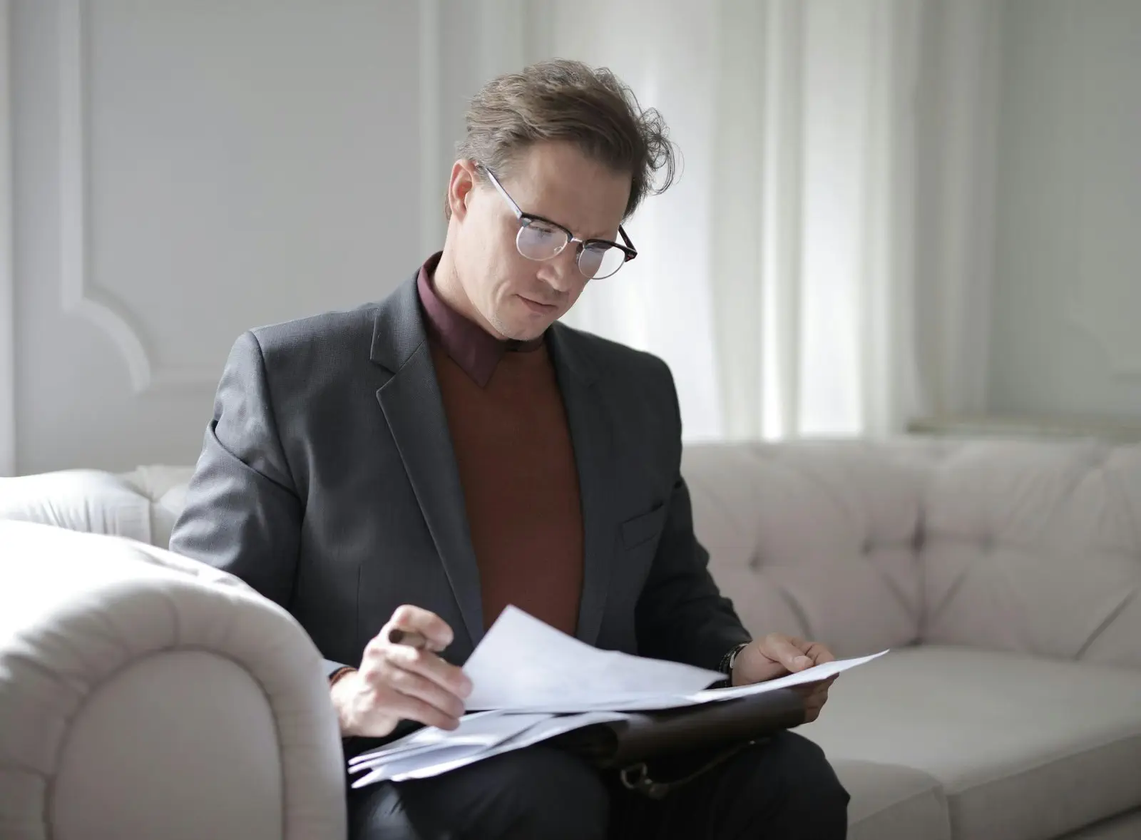 Man in suit with glasses reviews documents sitting on a beige couch in a bright room.