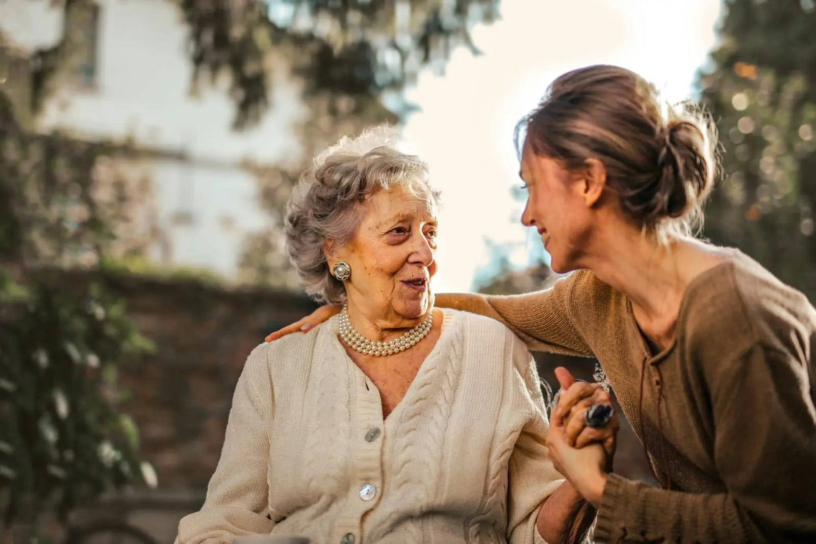 An elderly woman and a younger woman smile at each other while sitting outdoors, sharing a warm and affectionate moment.