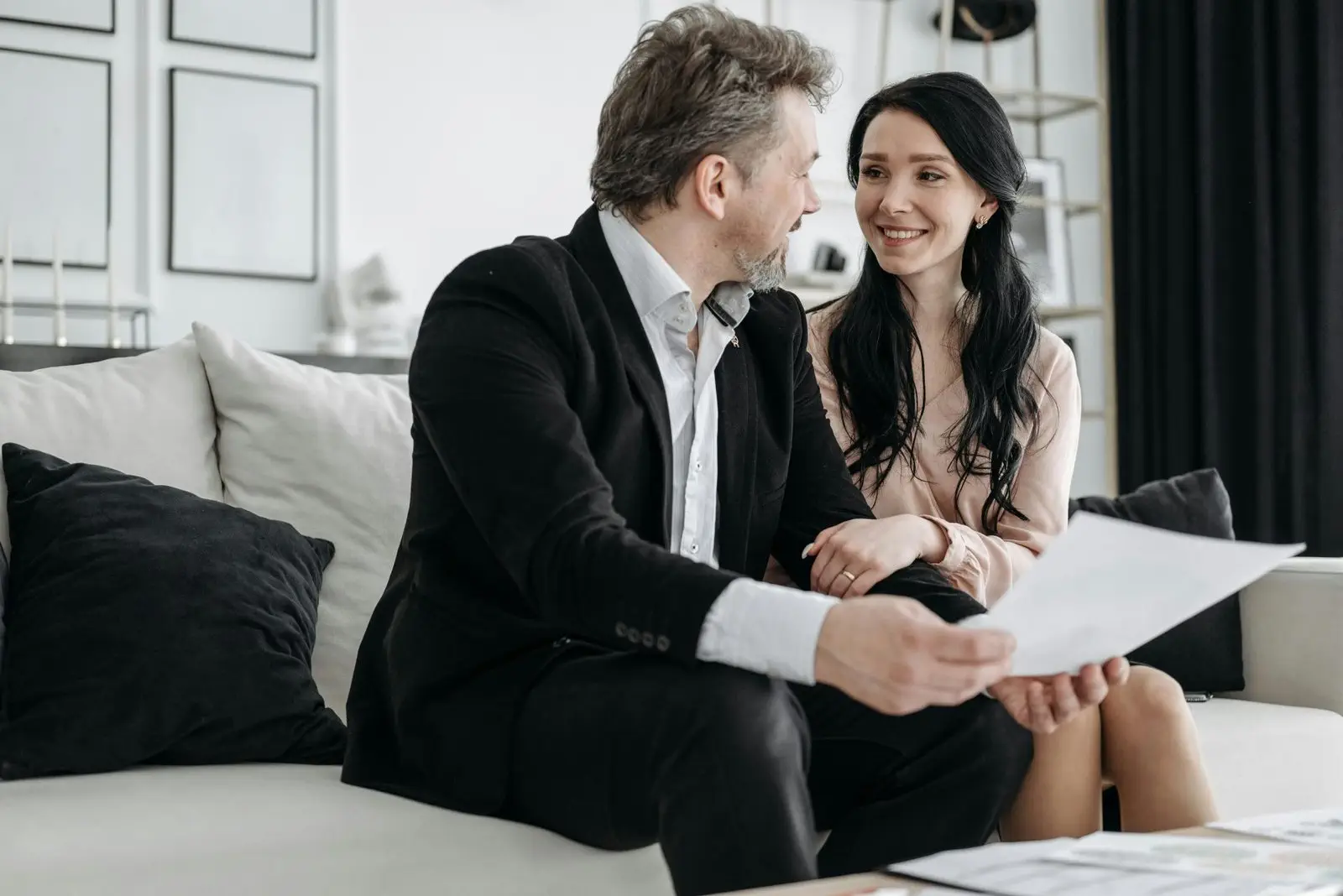 A man and a woman are sitting on a couch, smiling and holding a document, engaged in conversation in a modern living room.