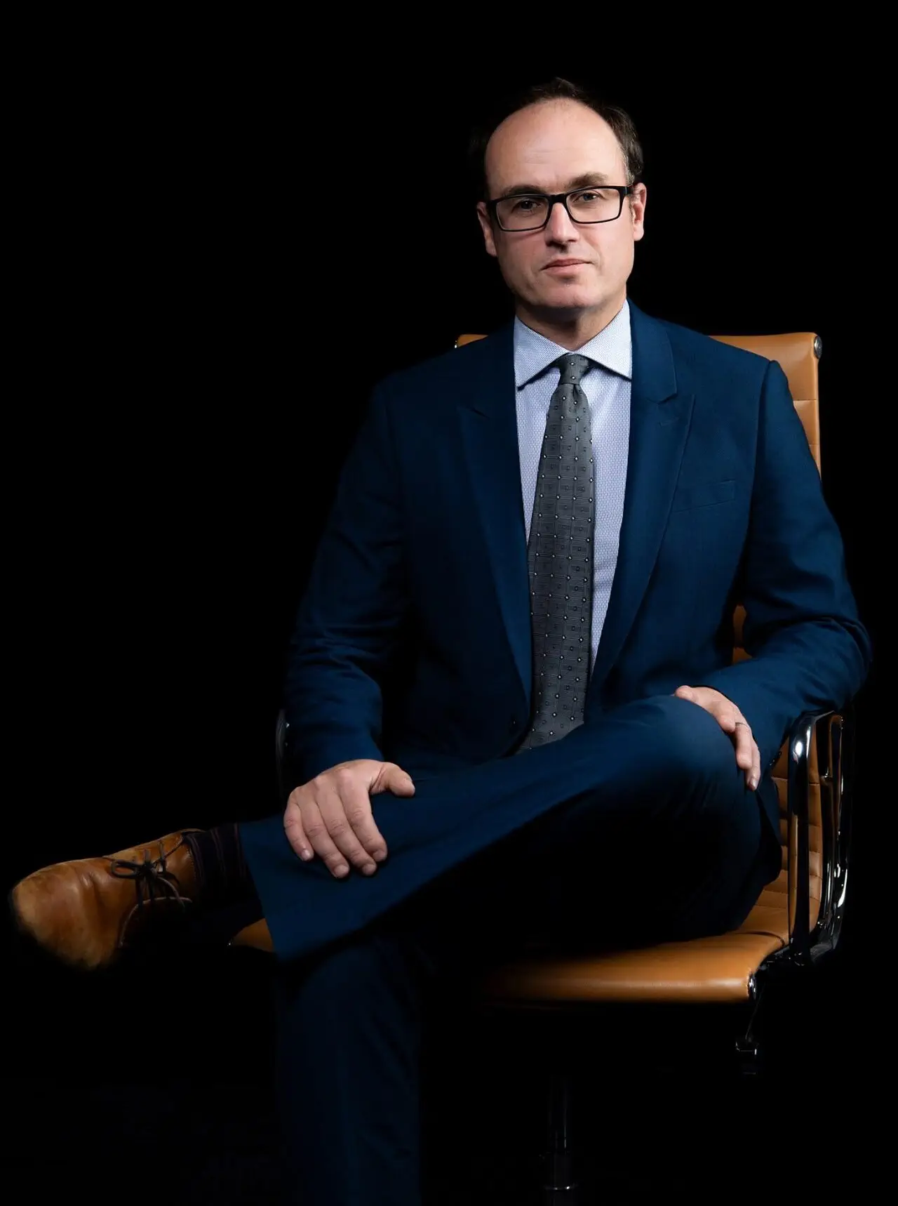 Iain Mark sits in an office chair, wearing a suit and glasses.