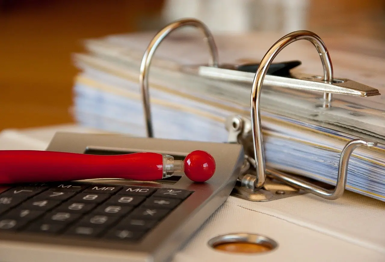 Calculator and red pen rest on a binder filled with financial documents, illustrating due diligence.